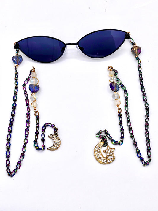 "Lunar Love" fashion eyewear by Orion Elise. Black-framed glasses with blue-tinted cat-eye lenses. Holographic drip and hearts, beads, and wire on armless chains. Gold gem-stoned moon and star charms symbolize cosmic enchantment. Illuminate your world with "Lunar Love" and shine brighter than ever before.
